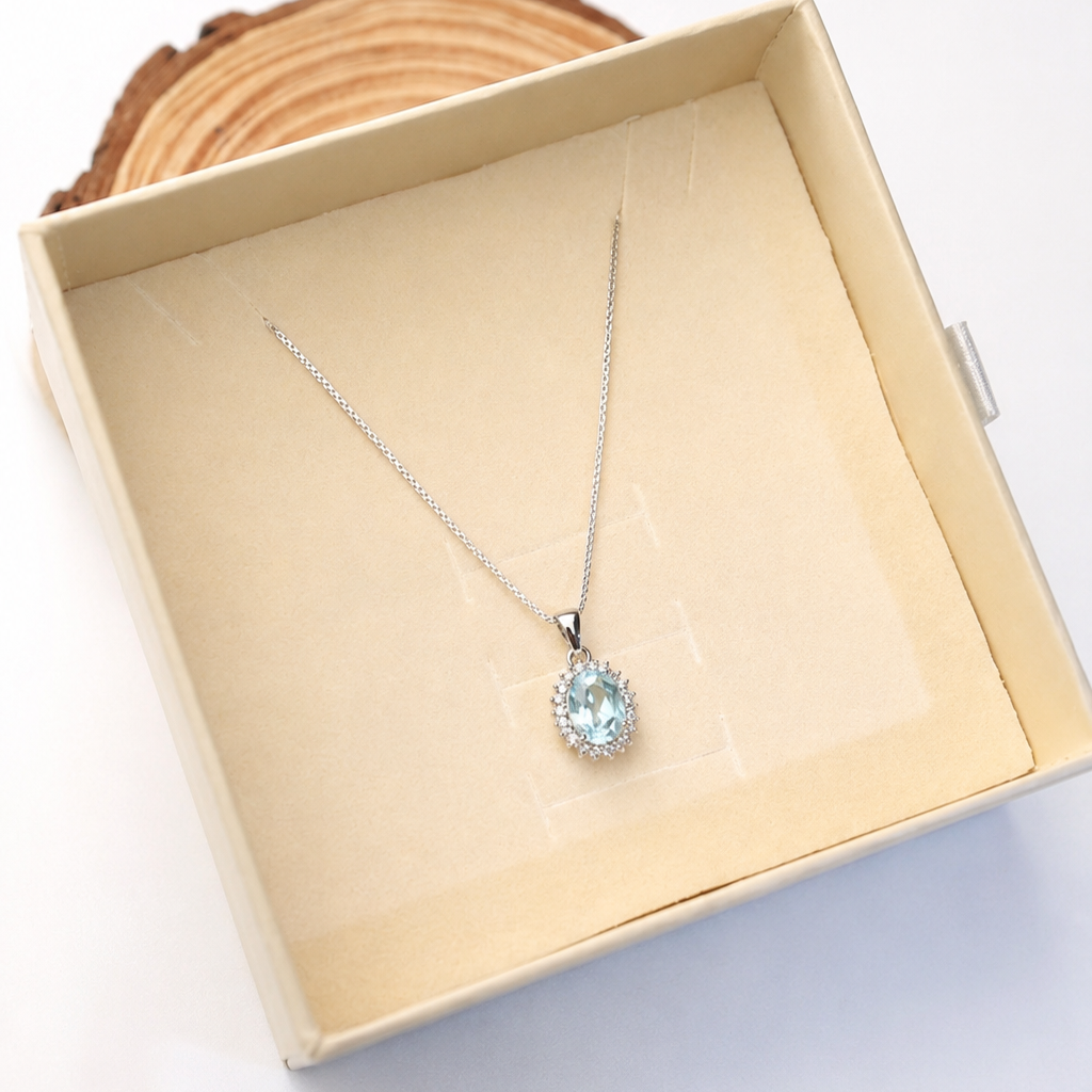 A silver pendant necklace with a light blue teardrop-shaped gemstone surrounded by small clear stones, displayed inside a beige jewelry box on a white background.