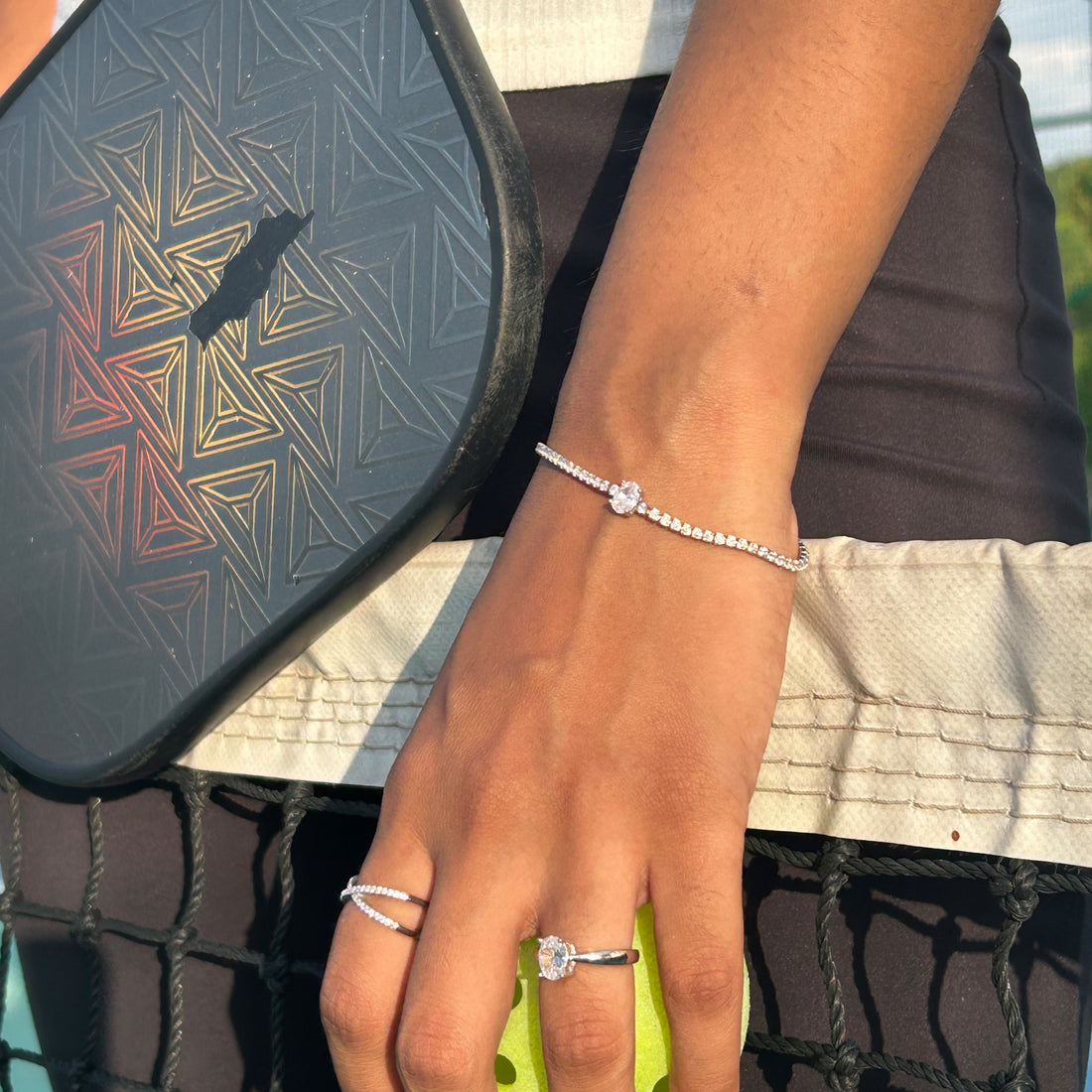 A close-up lifestyle shot of a person's hand and wrist wearing a delicate silver beaded bracelet with a small heart charm, alongside a tennis racket and tennis ball.