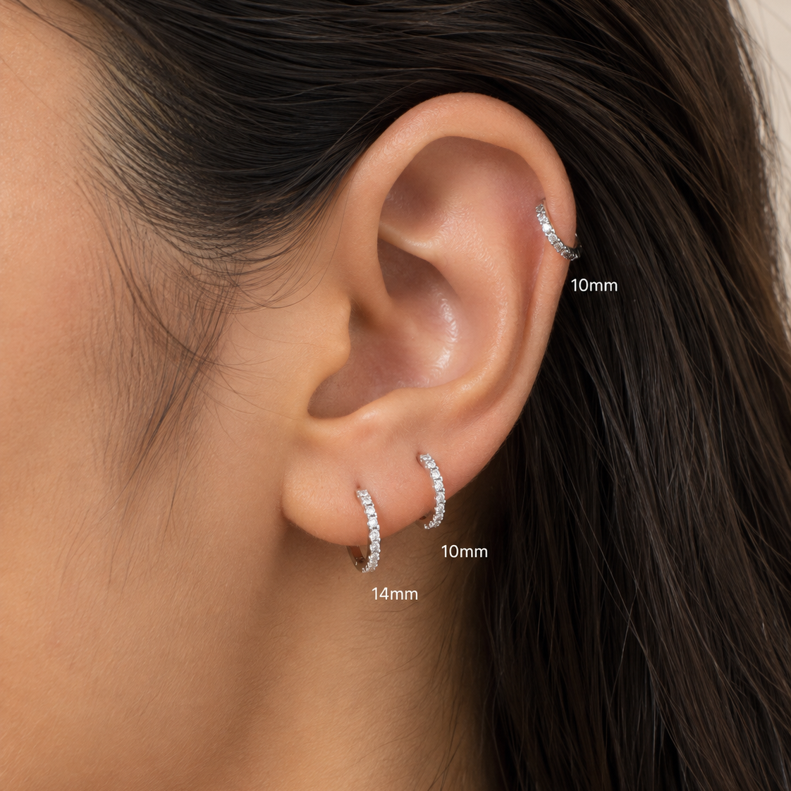 Close-up of a person's ear wearing three small silver hoop earrings at 10mm, 11mm, and 14mm, with size labels pointing to each earring.