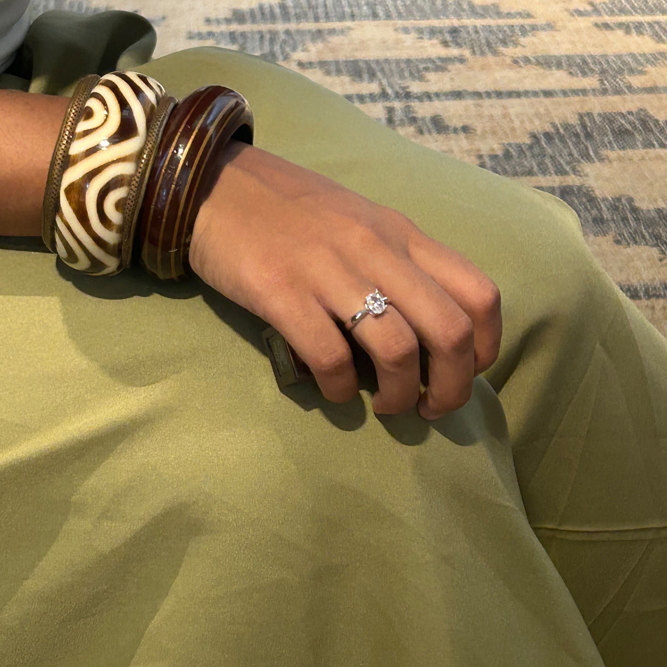 A close-up of a person's hand resting on their lap, wearing a silver-toned diamond engagement ring with a star-shaped center stone. The hand also has on a stack of brown and patterned wooden bangles, and the person is wearing an olive green skirt.