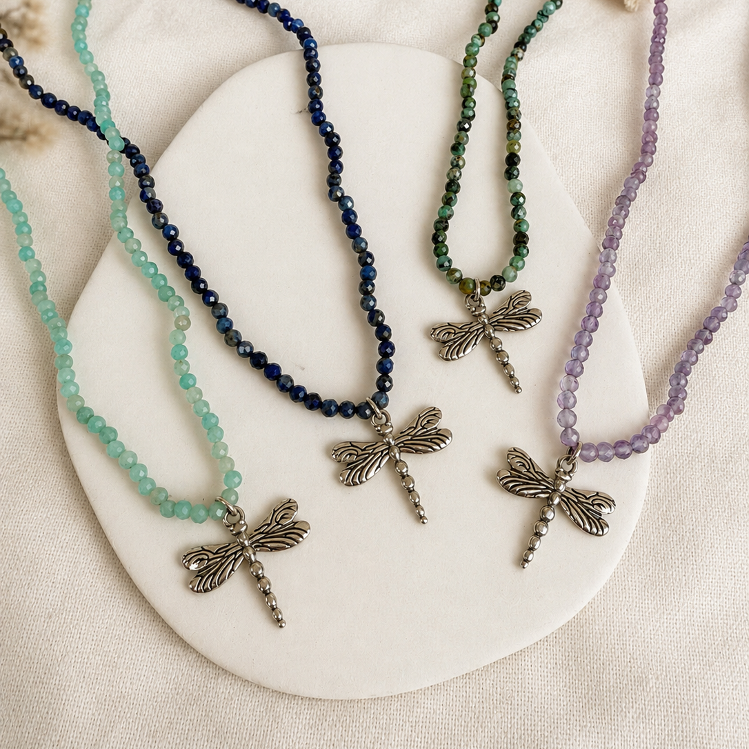 Three beaded dragonfly necklaces in mint green, dark blue, and lavender purple, displayed on a white oval tray over a textured beige fabric background.