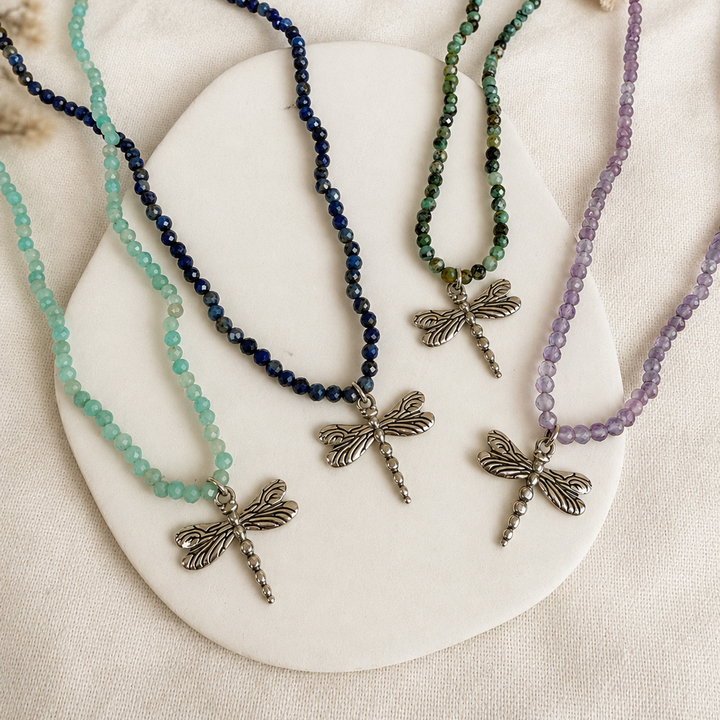 Three beaded dragonfly necklaces in mint green, dark blue, and lavender purple, displayed on a white oval tray over a textured beige fabric background.