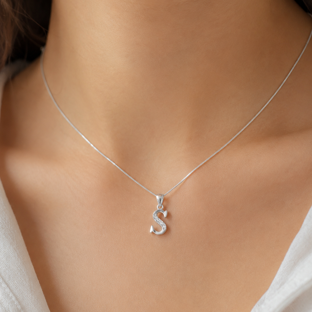 A close-up of a model's neck and chest wearing a delicate silver chain necklace with a small silver initial pendant spelling 'S'.