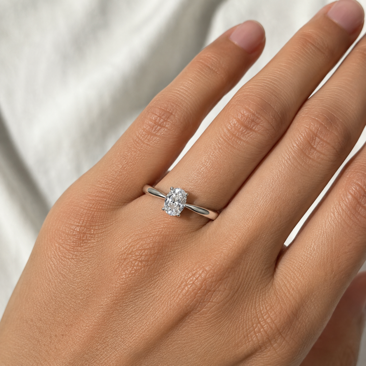 A close-up of a hand wearing a silver solitaire ring with an oval-cut clear gemstone, set against a soft white fabric background.