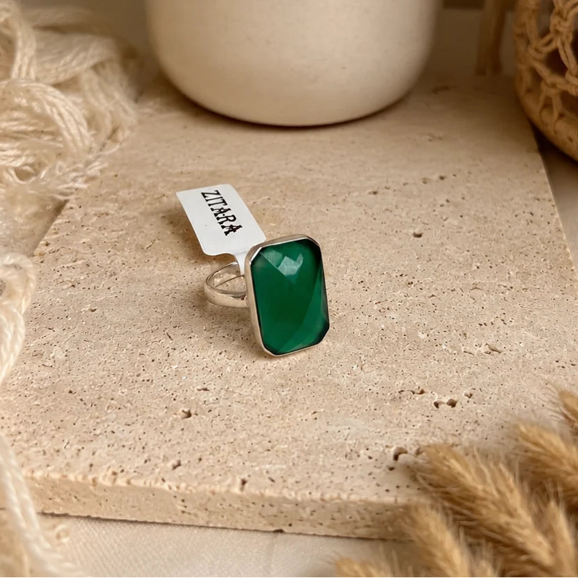 A silver ring with a rectangular green stone, displayed on a textured beige stone slab with a small paper tag that reads 'LIRA'. The background features neutral-toned decorative props including a cream ceramic vase, dried flowers, and a woven basket.