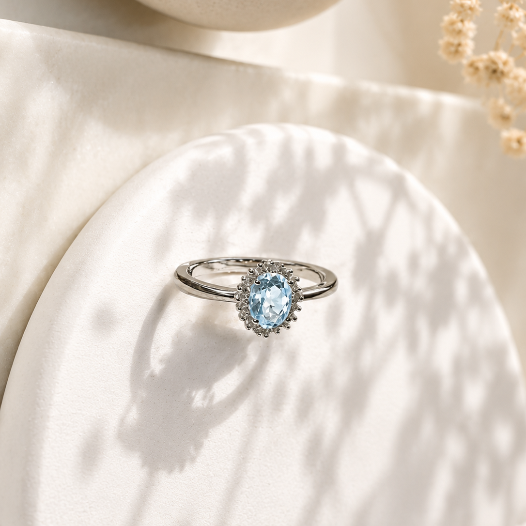A silver ring with a light blue oval gemstone and small clear accent stones, displayed on a white textured pedestal with soft shadows and dried flowers in the background.