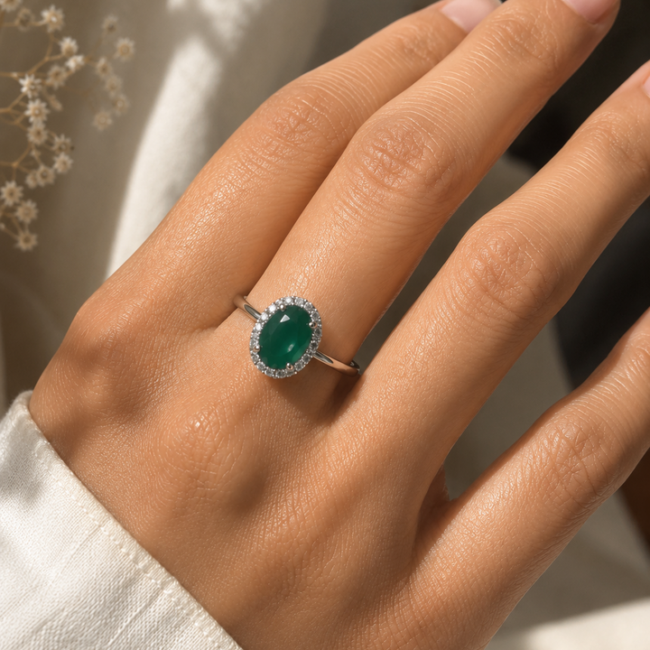 A close-up of a hand wearing a silver ring with an oval green gemstone surrounded by a halo of small clear stones, set against a soft neutral background.