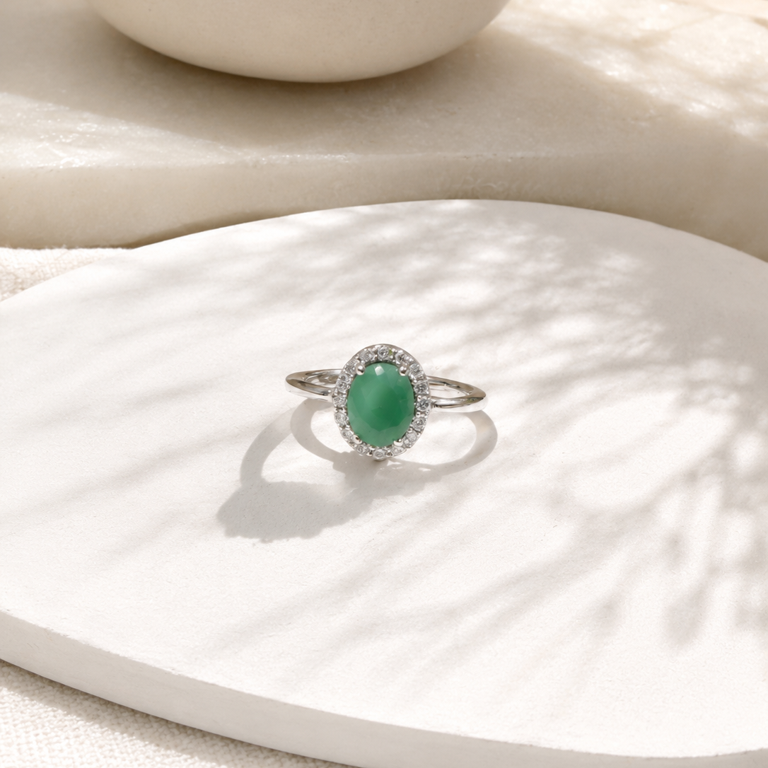 A silver ring featuring an oval green jade-like center stone surrounded by a halo of small clear stones, displayed on a white sculptural surface with soft shadows.