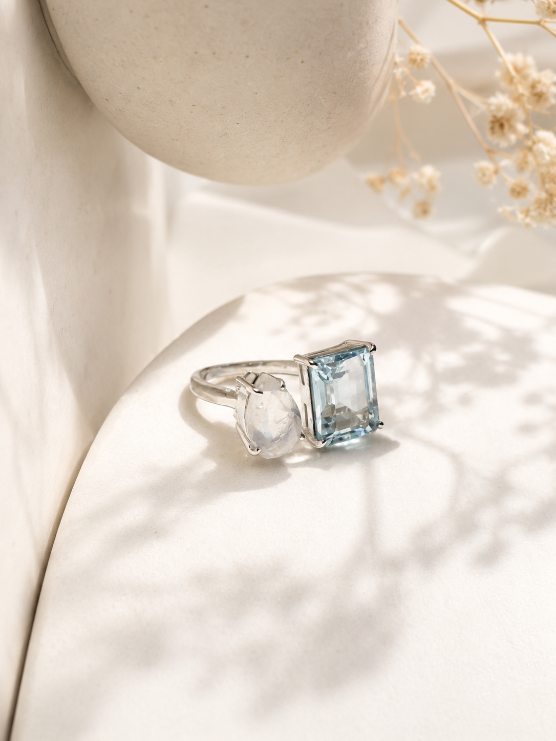 A silver ring featuring a rectangular light blue gemstone and a smaller round clear stone, displayed on a white curved surface with soft shadows and dried flowers in the background.