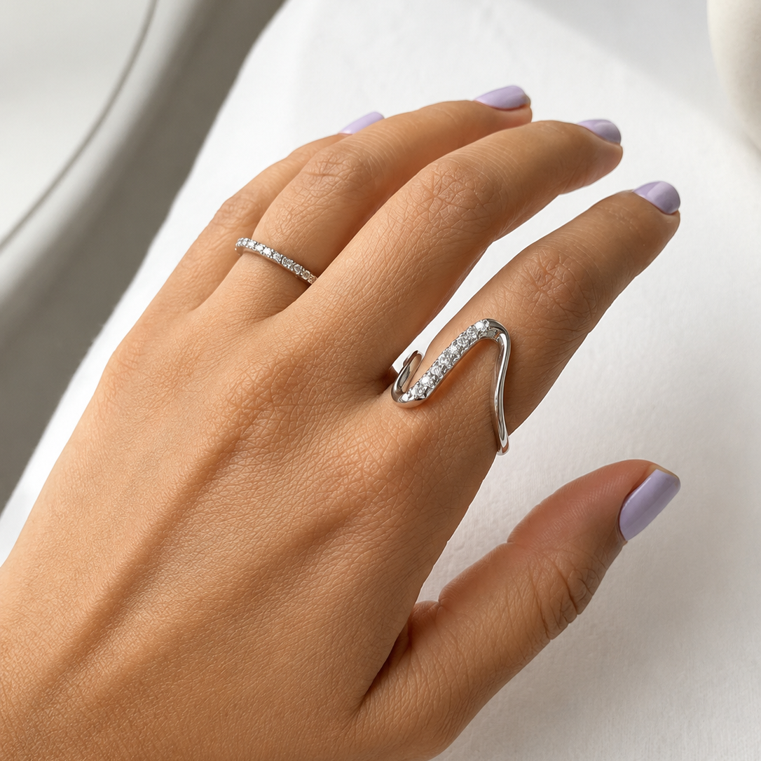 A close-up of a hand with lavender nail polish wearing a silver wavy ring with pavé-set clear stones, resting on a white textured surface.