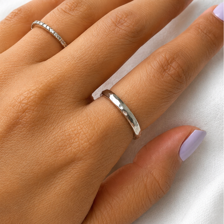 A close-up of a hand with lavender nail polish wearing two silver rings, one a delicate pavé band and the other a smooth hammered band, resting on a soft white fabric background.