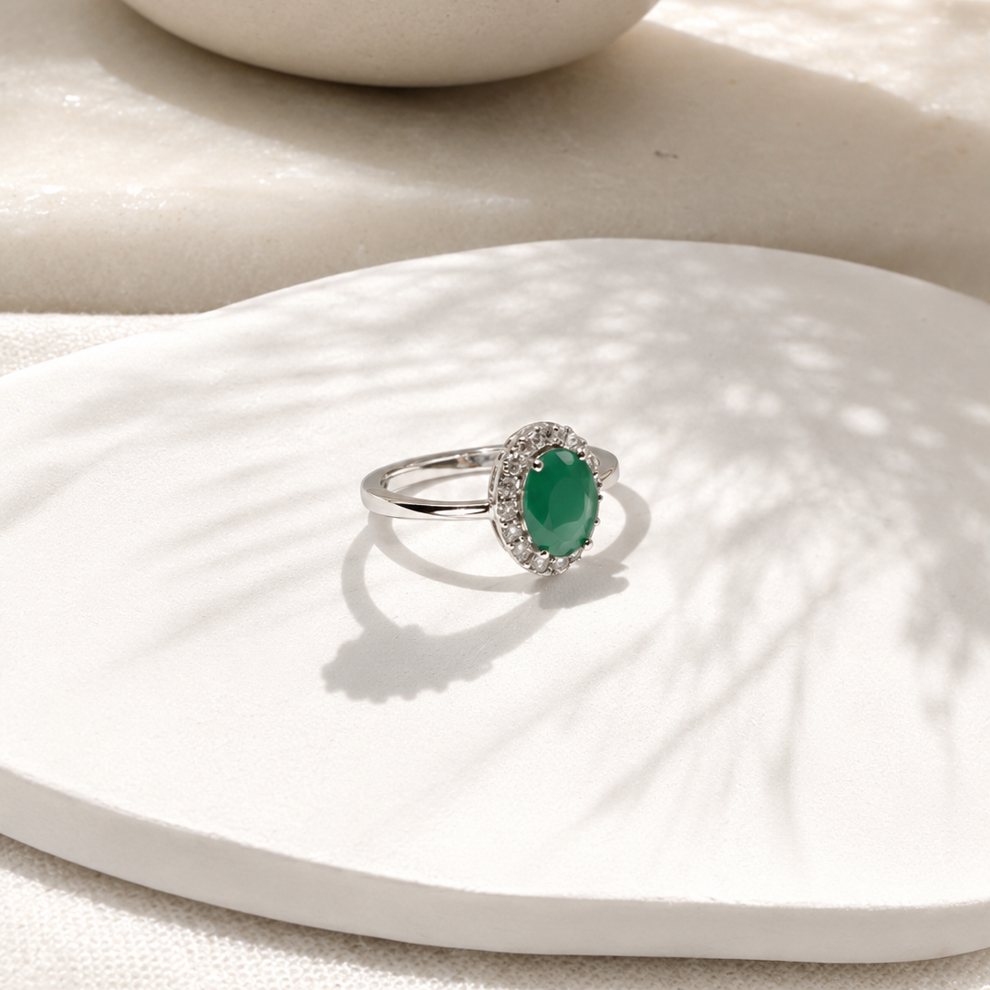 A silver ring with an oval green gemstone surrounded by small clear stones, displayed on a white textured surface with soft shadows.