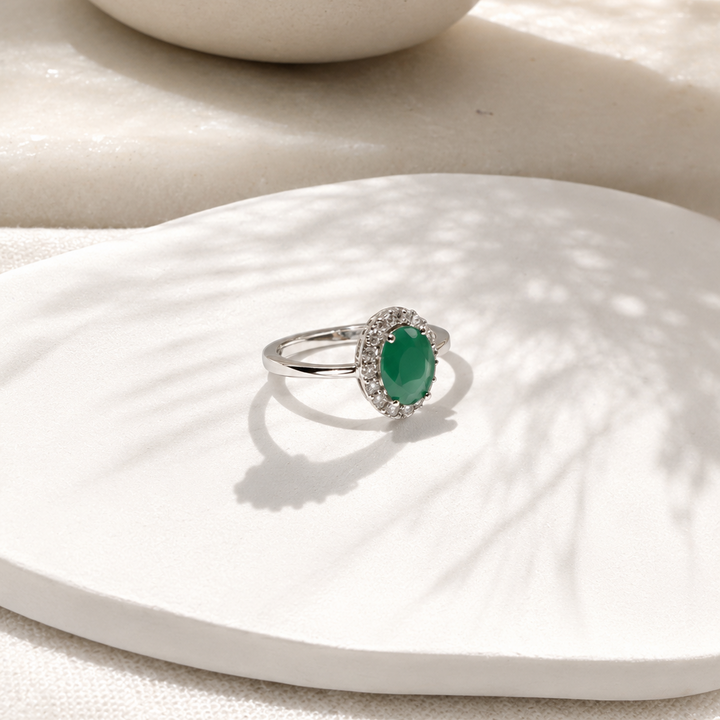A silver ring with an oval green gemstone surrounded by small clear stones, displayed on a white textured surface with soft shadows.