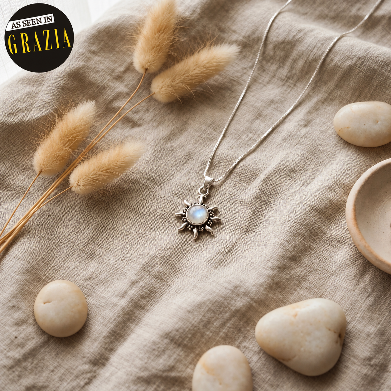A silver-toned necklace with a sunburst pendant featuring a round iridescent moonstone, styled on a beige linen fabric with dried bunny tail grass, smooth stones, and a small wooden dish. A black circular 'AS SEEN IN GRAZIA' badge is in the top-left corner.