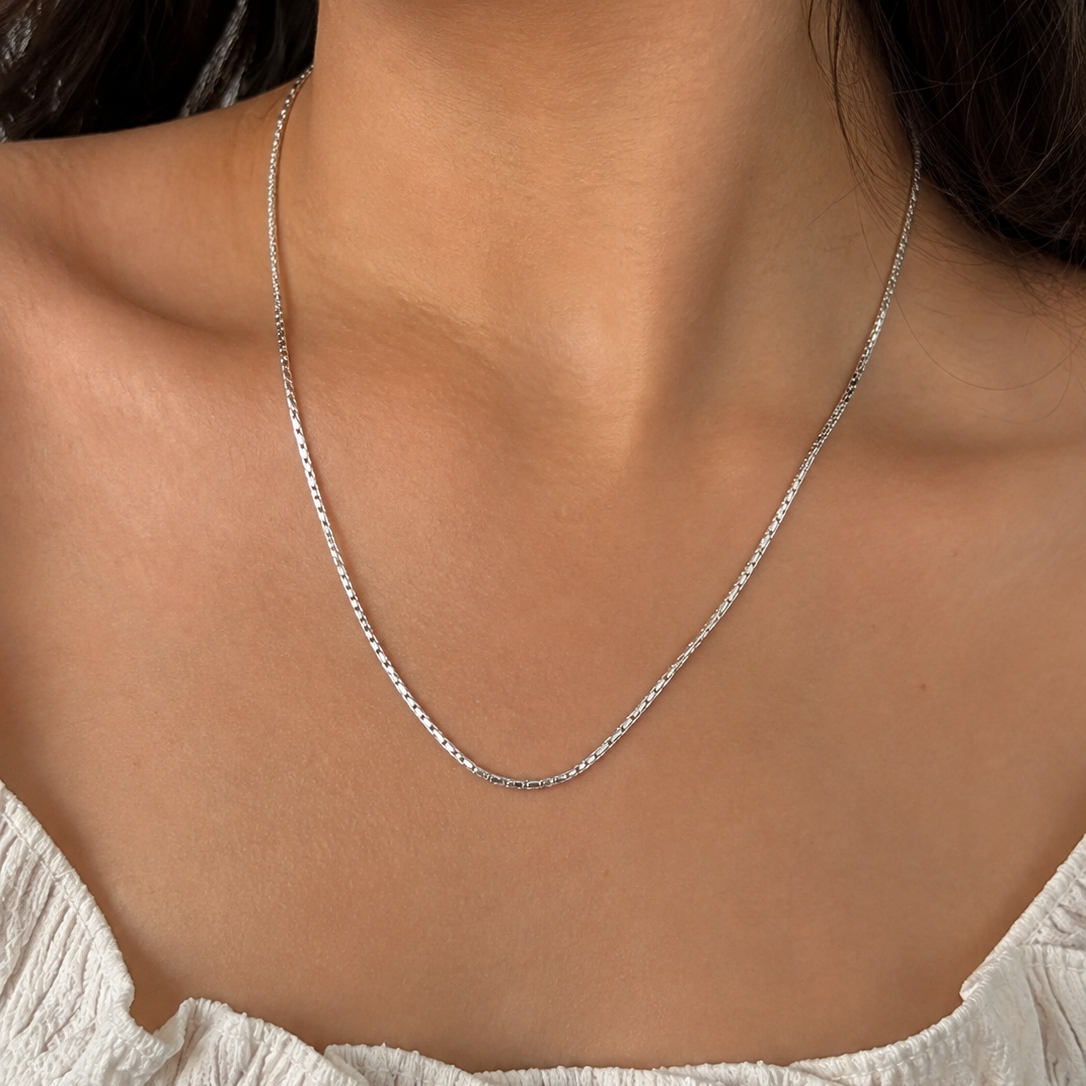 A close-up of a model's neck and upper chest wearing a delicate silver chain necklace with a small pendant.