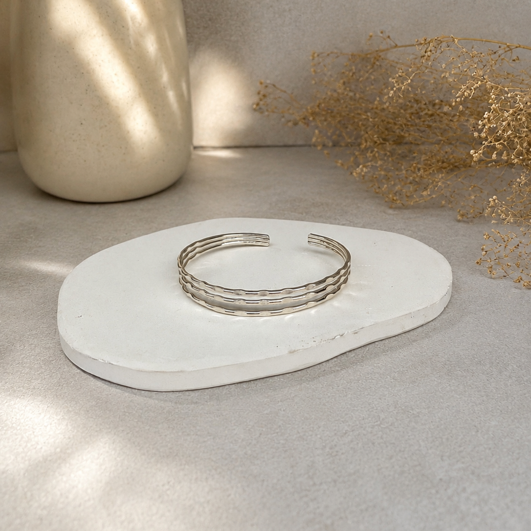 A stack of three textured silver cuff bracelets displayed on a small white ceramic dish, set against a neutral beige background with a ceramic vase and dried botanicals.