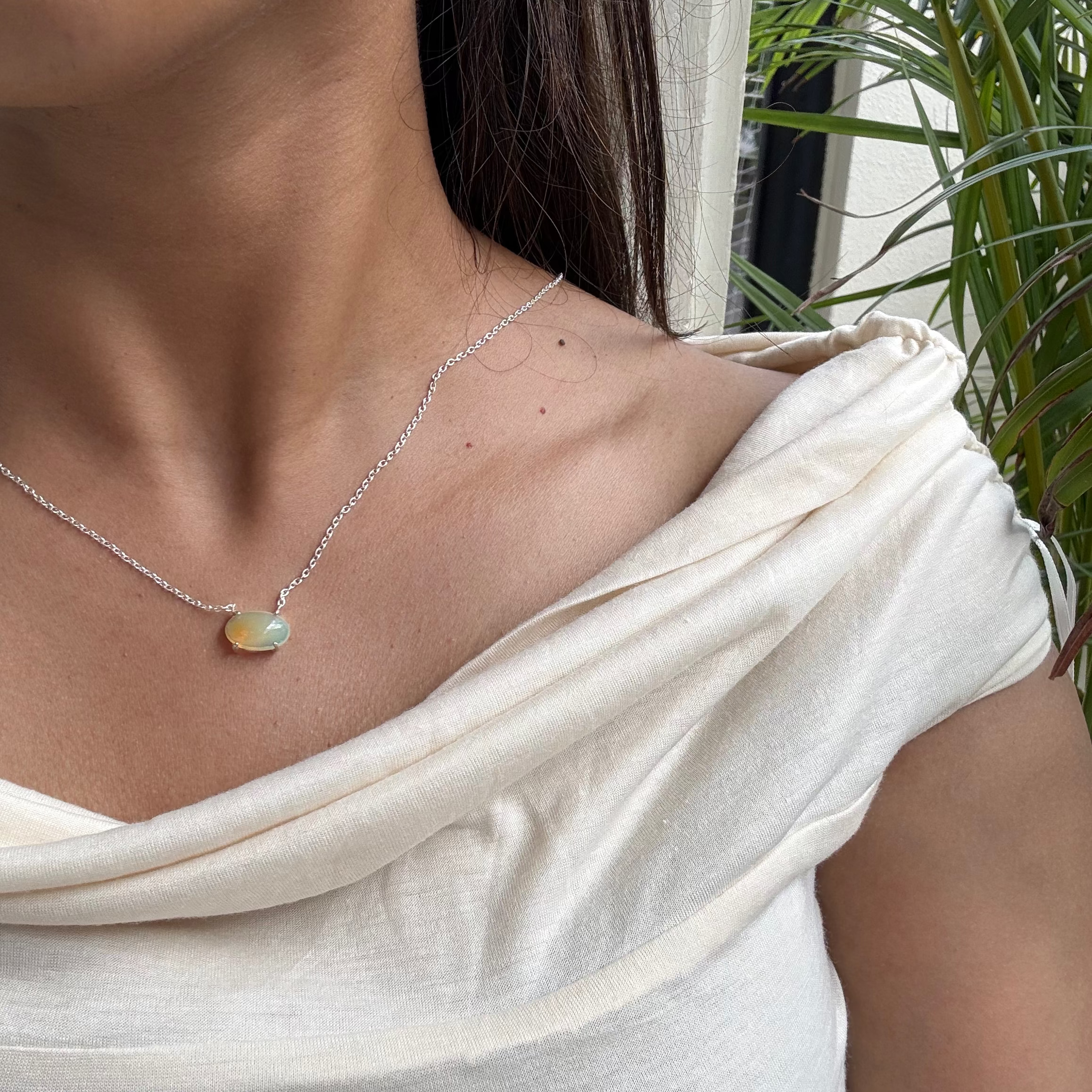 Woman wearing a silver necklace with a pendant, standing outdoors with plants in the background.