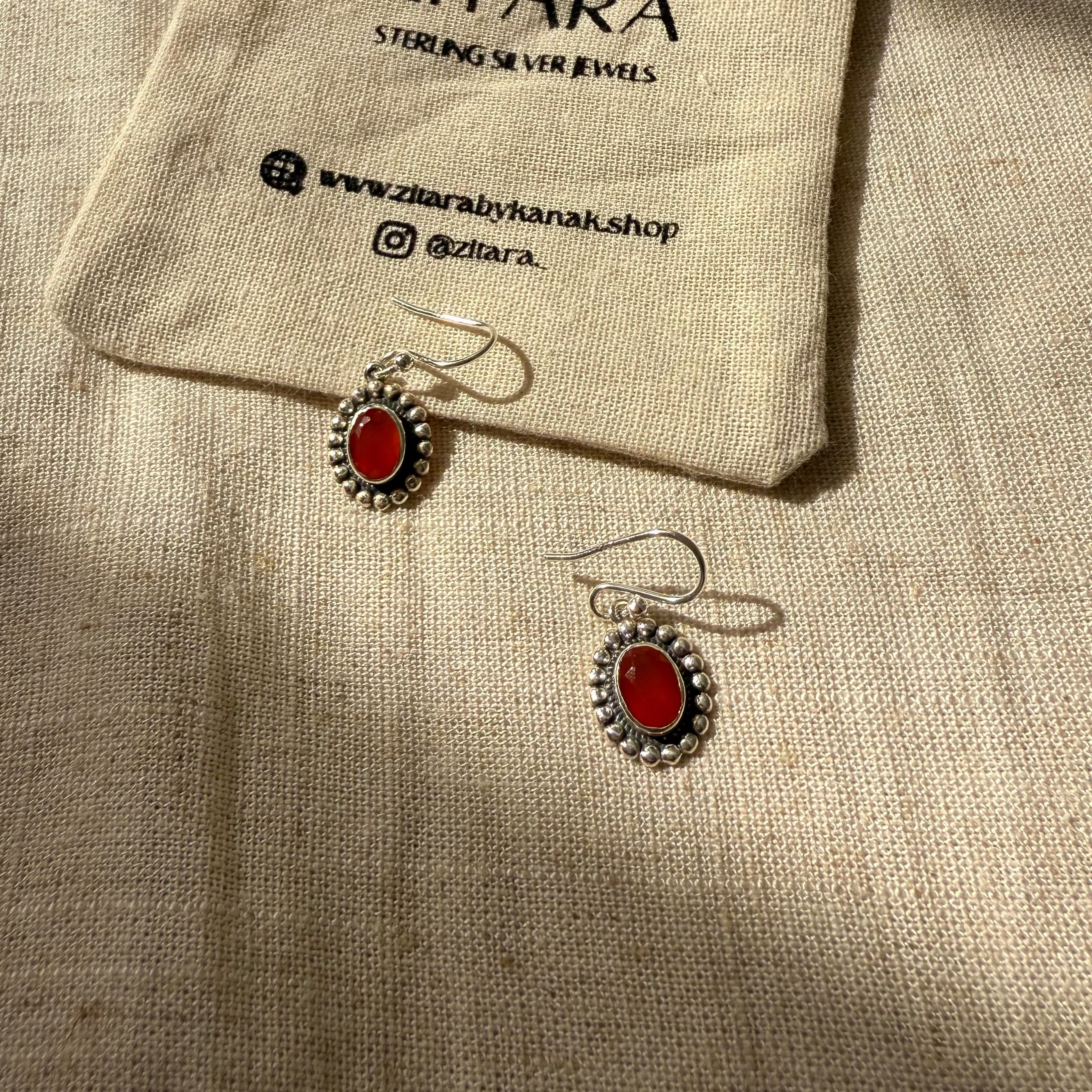 A pair of oval drop earrings with red stones and silver settings, displayed on a beige linen fabric next to a small branded drawstring pouch that reads ZITARA.