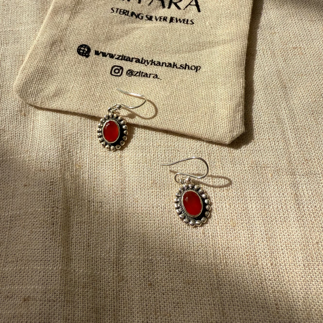 A pair of oval drop earrings with red stones and silver settings, displayed on a beige linen fabric next to a small branded drawstring pouch that reads ZITARA.