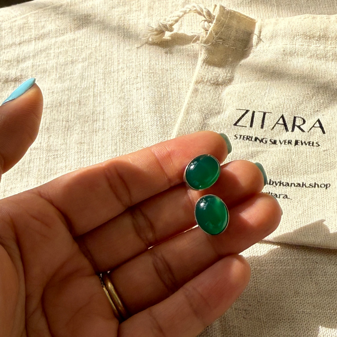 A close-up of a hand holding two oval green cabochon stud earrings with silver settings, positioned in front of a white branded fabric bag that reads 'ZITARA Sterling Silver Jewels'.
