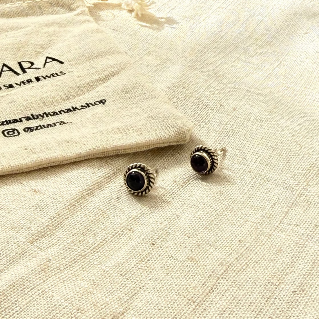 A pair of silver-toned stud earrings with black circular centers, displayed on a textured cream linen surface next to a branded cream drawstring pouch labeled ZITARA.
