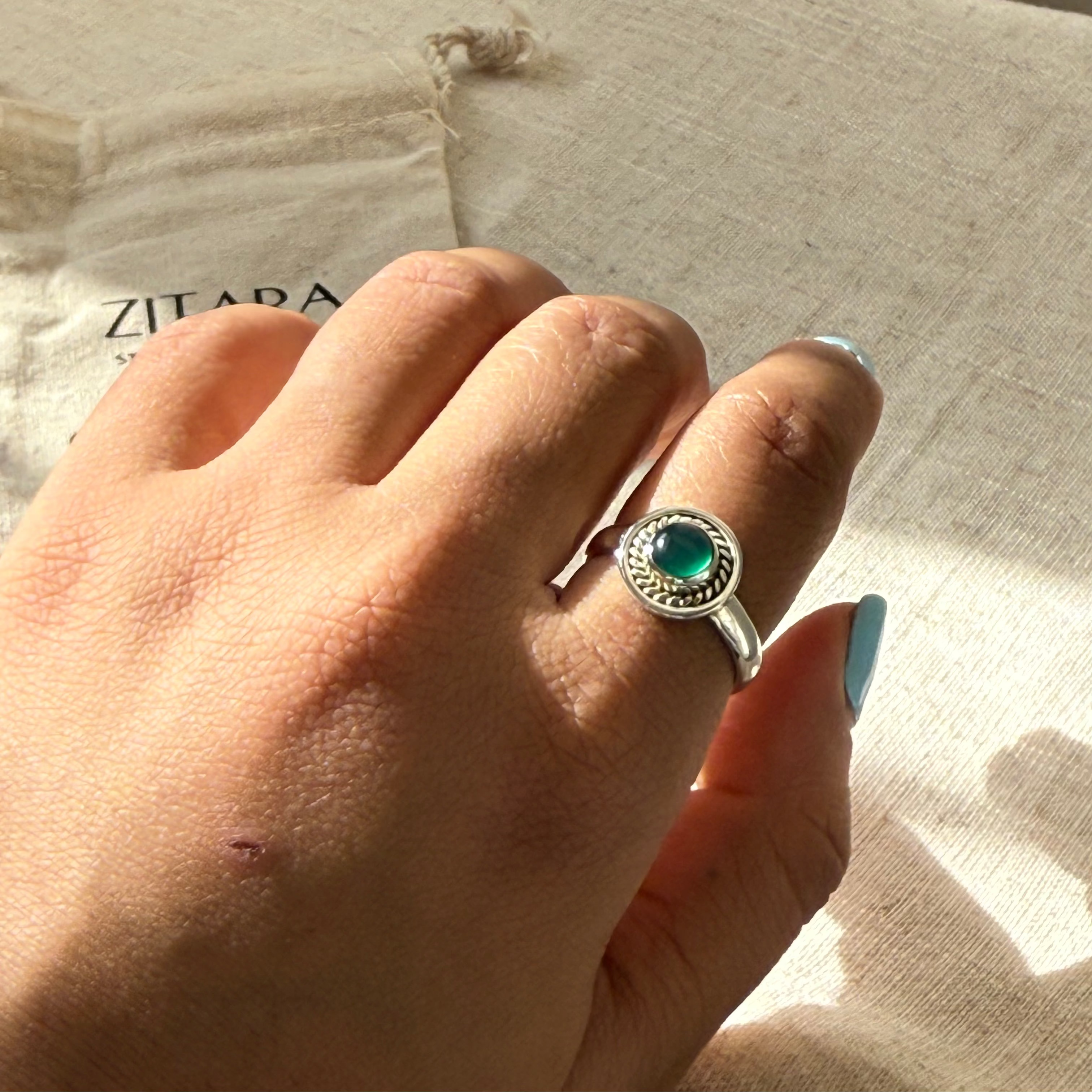 A hand wearing a silver ring with a round green stone, resting on a beige linen surface with sunlight and shadows.