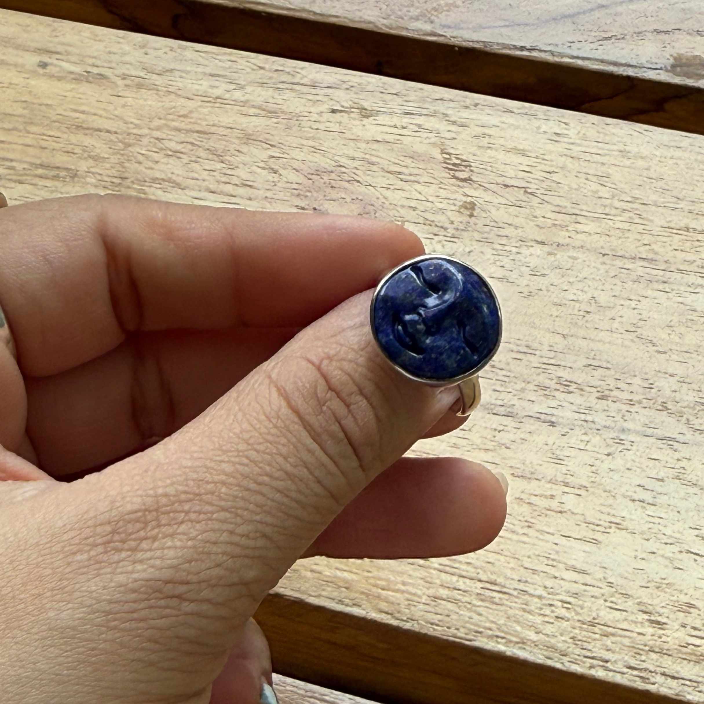 Hand holding a blue stone ring with other rings on a wooden surface