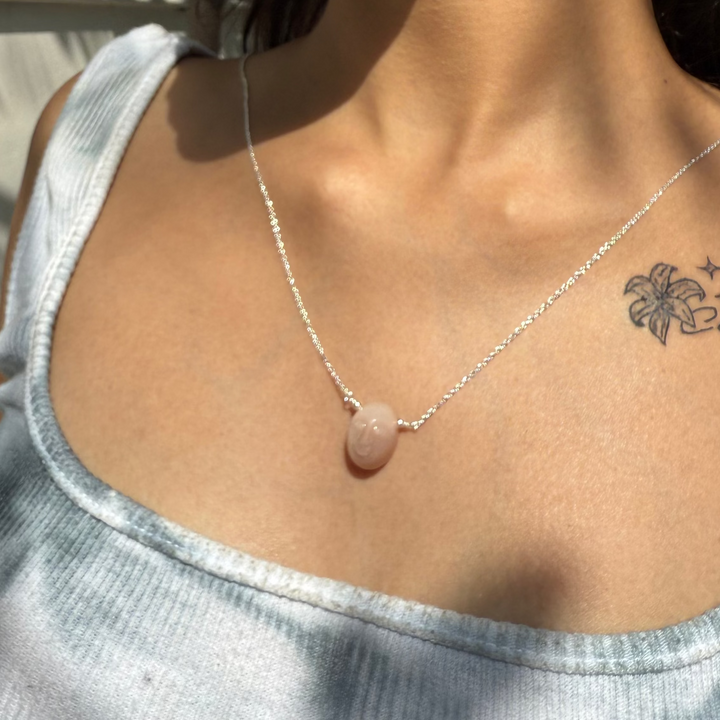 Close-up of a person wearing a necklace with a pink stone and earrings, with a neutral background.