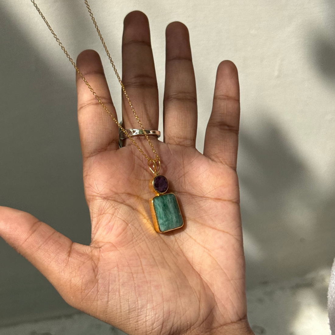Hand holding a gold necklace with a green pendant against a neutral background