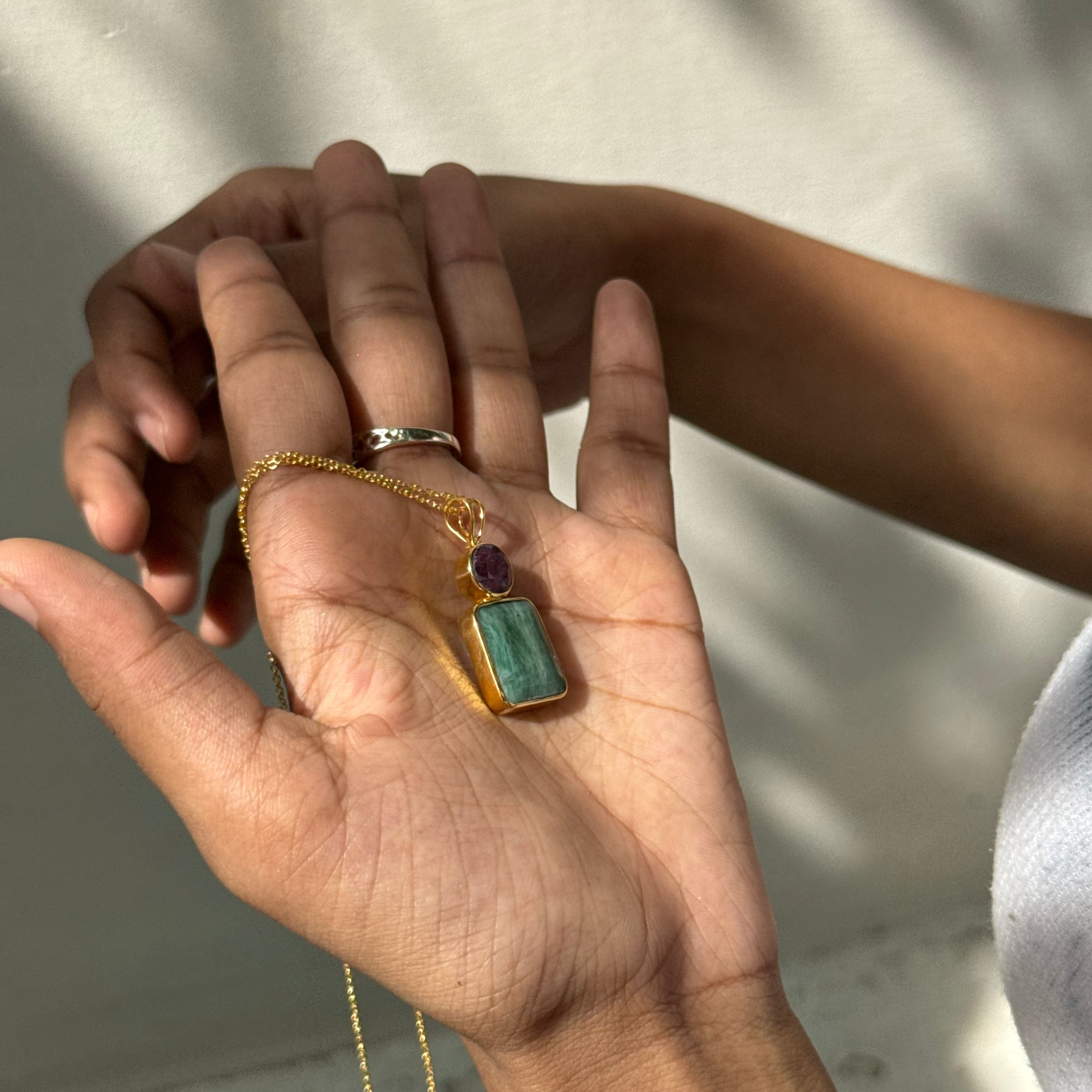 Hand holding a gold necklace with a green pendant against a neutral background