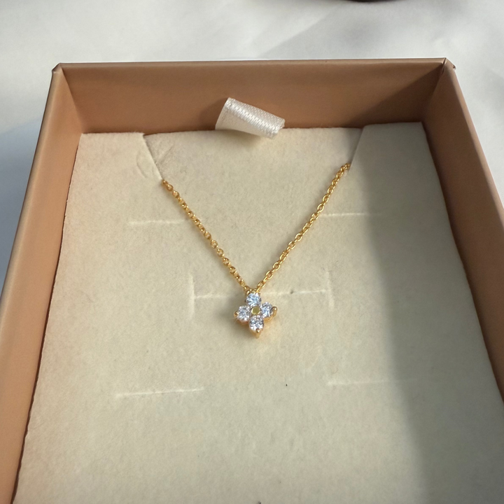 Gold necklace with a floral pendant in a box on a white surface with a dark brown vase.
