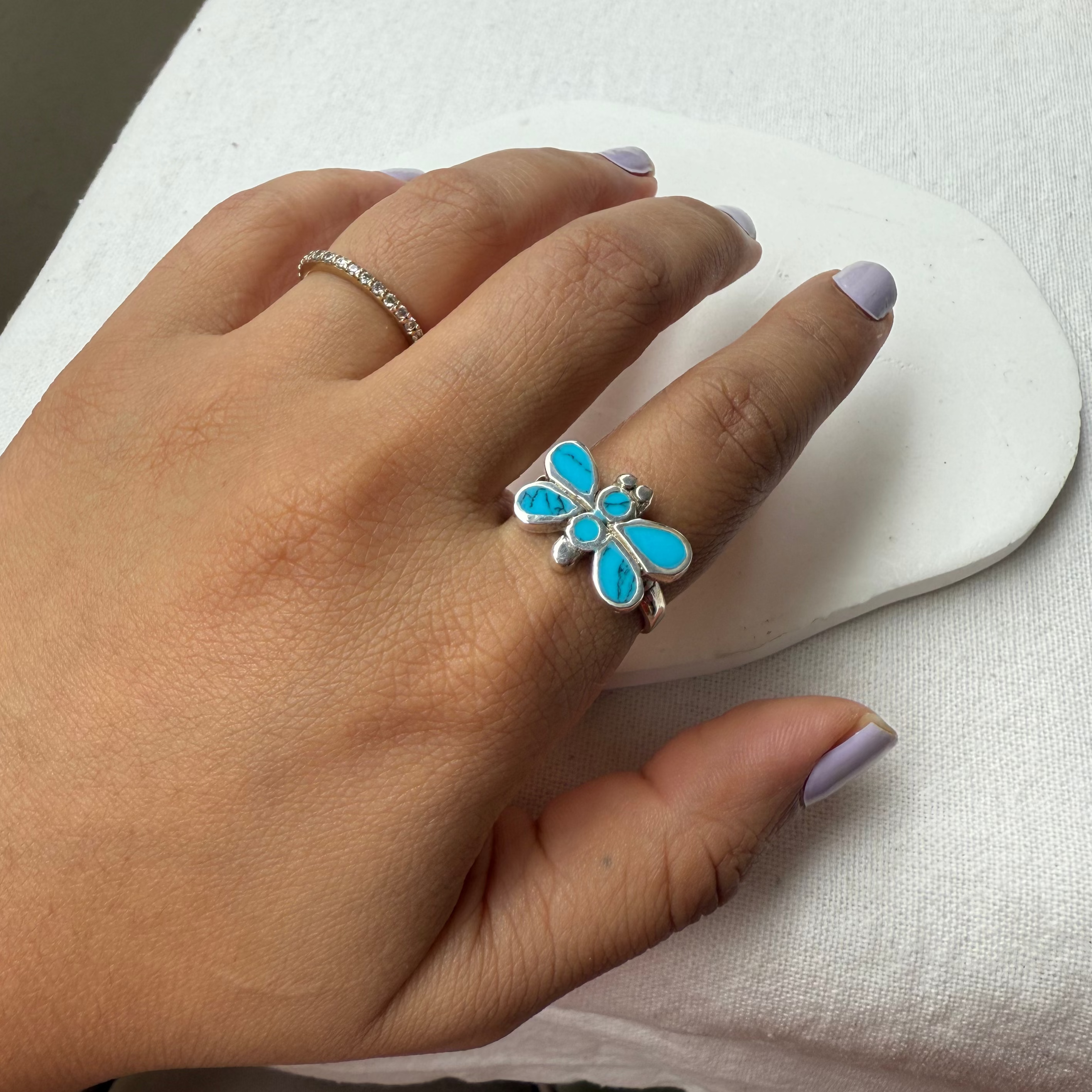 A hand wearing a silver ring with a butterfly-shaped turquoise inlay, resting on a white surface.
