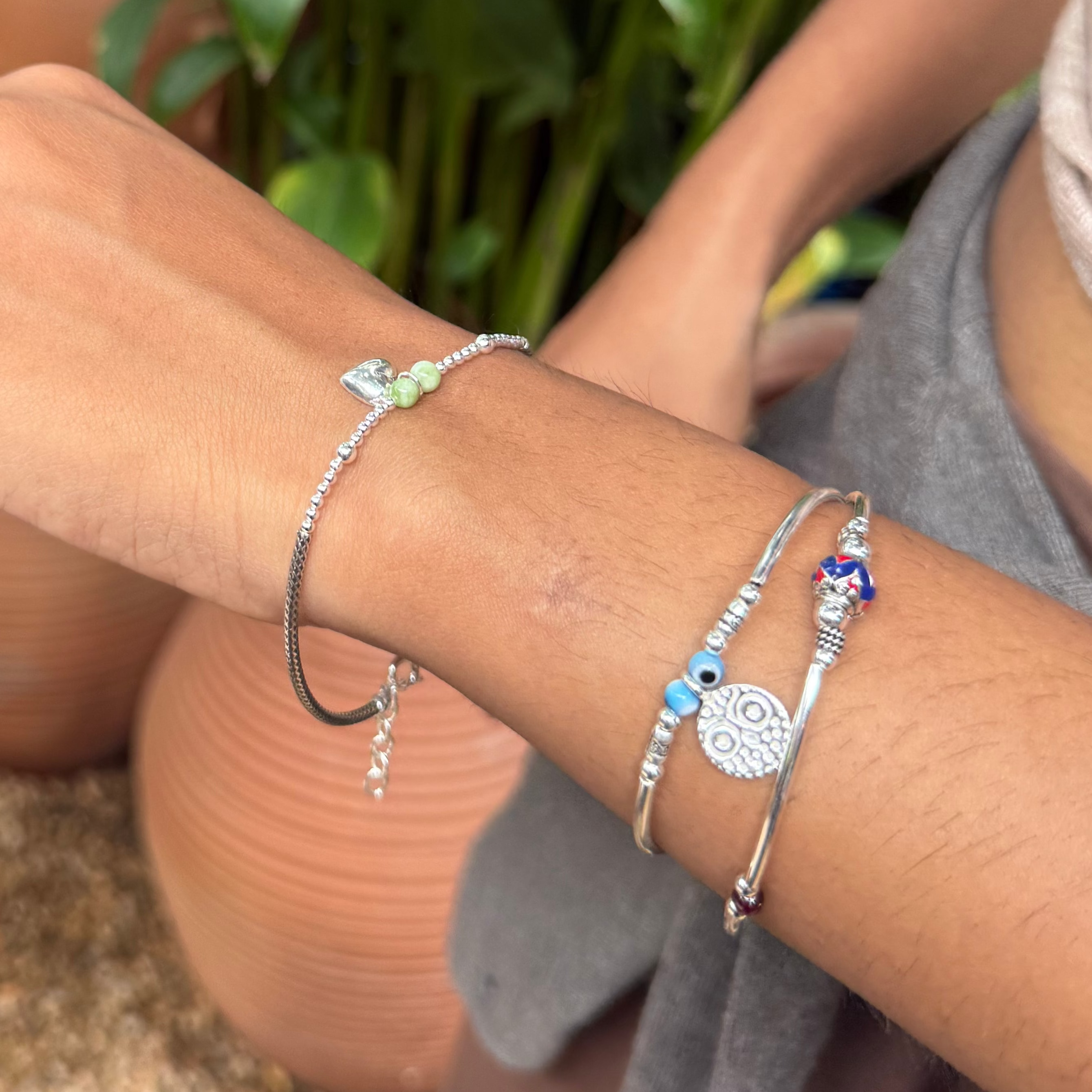 A close-up of a person's wrist and hand wearing multiple delicate silver bracelets, including a bangle with a small green leaf charm and others with blue and silver details, set against a background of potted green plants and terracotta pots.