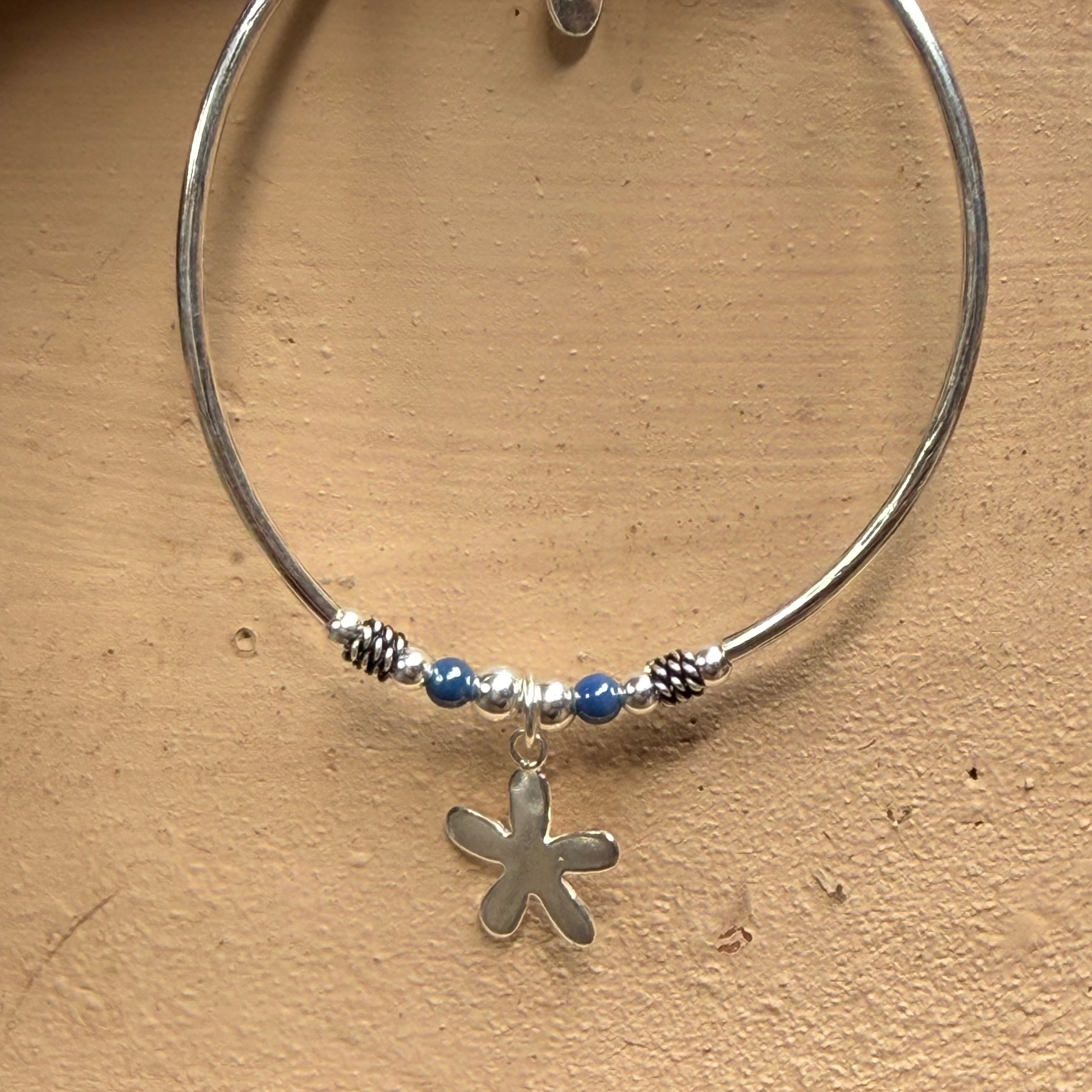 A silver-toned circular bangle bracelet with a small flower-shaped charm, blue beads, and textured spacer details, displayed against a textured beige background.