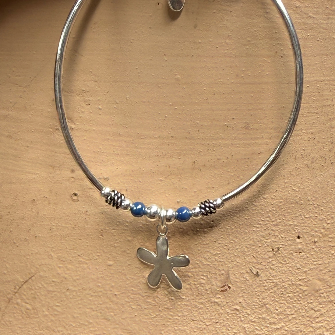 A silver-toned circular bangle bracelet with a small flower-shaped charm, blue beads, and textured spacer details, displayed against a textured beige background.