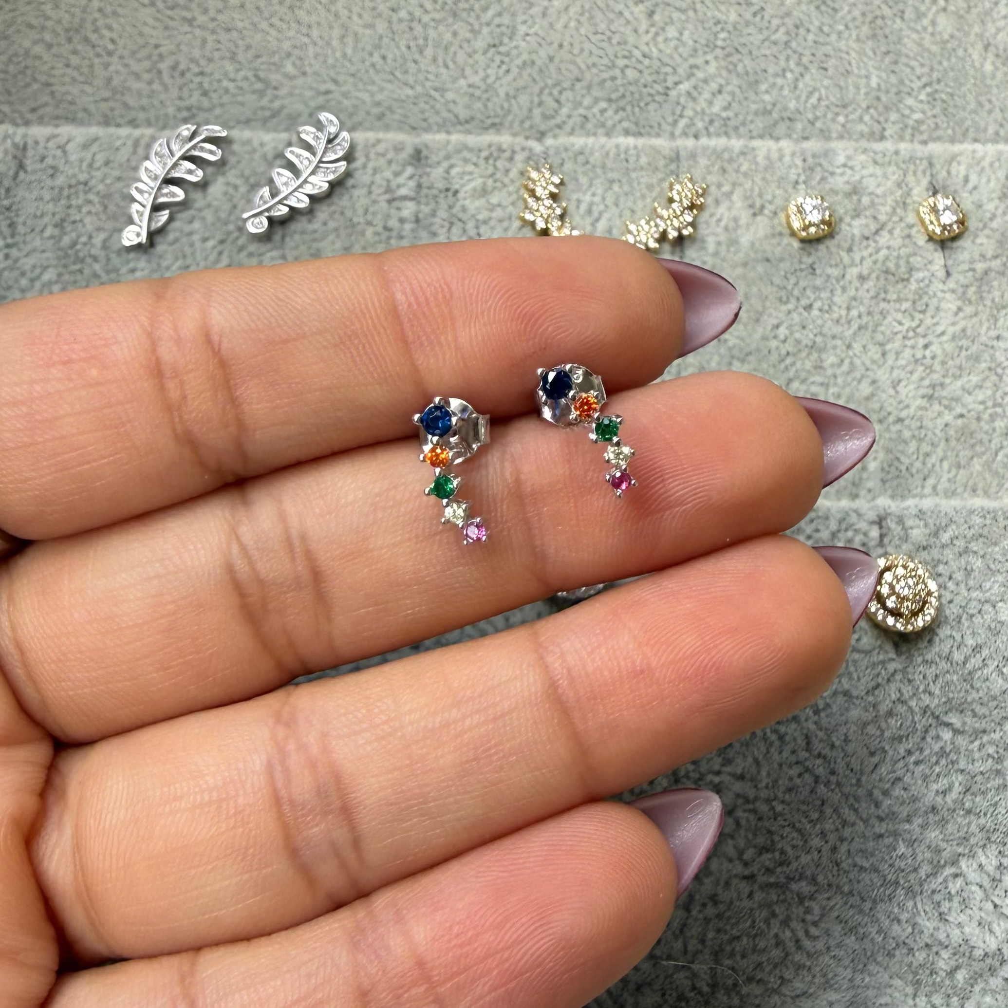 A hand holding a pair of small dangling earrings with blue, pink, and green stones, with other jewelry displayed on a gray velvet tray in the background.