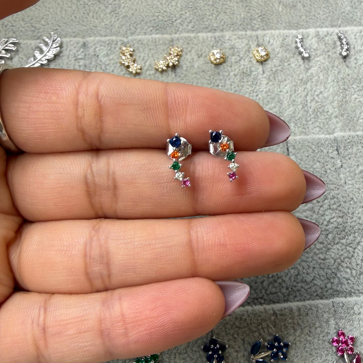 A hand with lavender nail polish holds a pair of small silver crescent-shaped hoop earrings decorated with colorful gemstones, with other jewelry pieces displayed on a gray velvet jewelry tray in the background.
