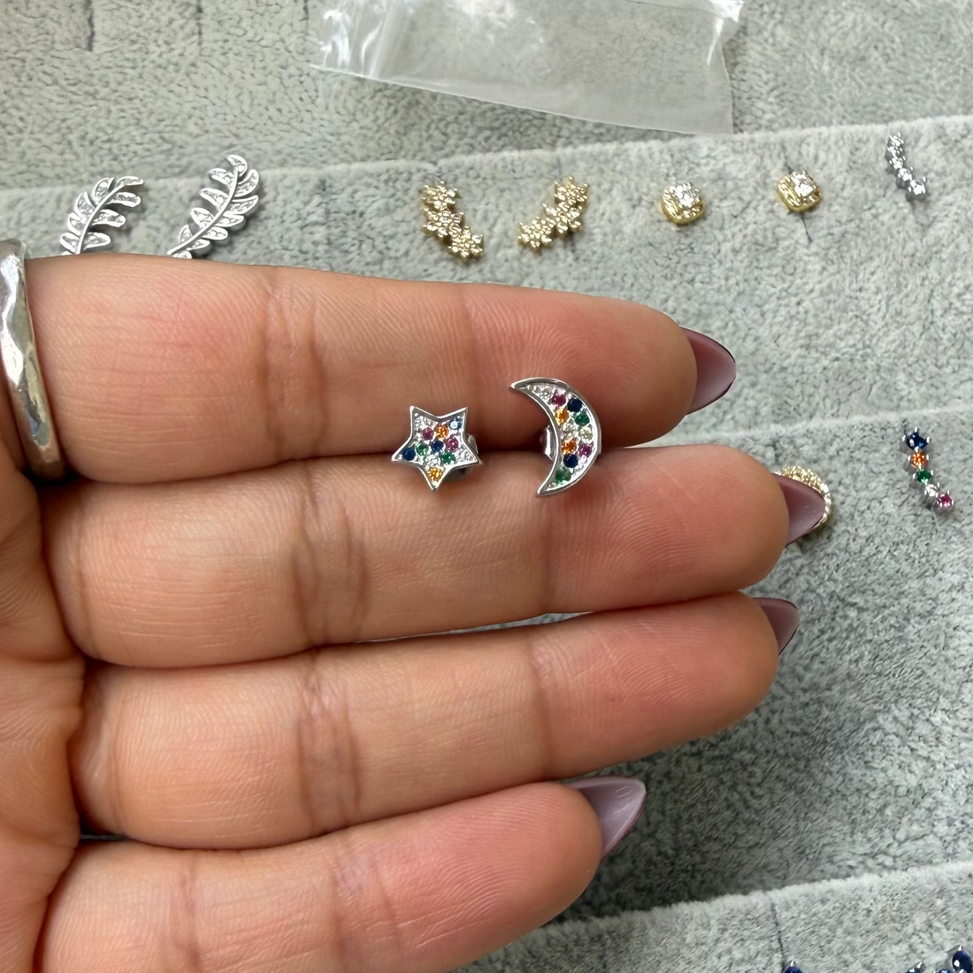 A hand holding a pair of small silver crescent moon and star stud earrings with colorful rhinestones, displayed over a gray jewelry tray with other earrings.