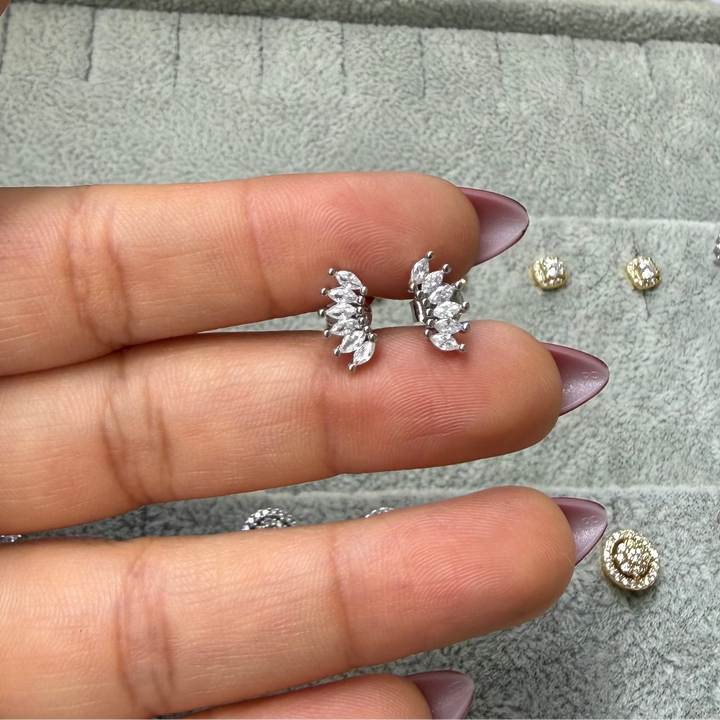 A pair of silver-toned leaf-shaped stud earrings with clear stones, held between fingers over a gray jewelry tray with other earrings nearby.