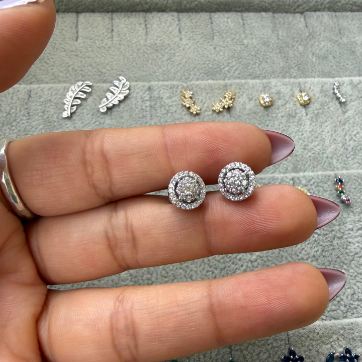 A hand with lavender-painted nails holds a pair of round silver stud earrings with clear pavé-set stones, positioned over a gray jewelry display tray containing other earrings.
