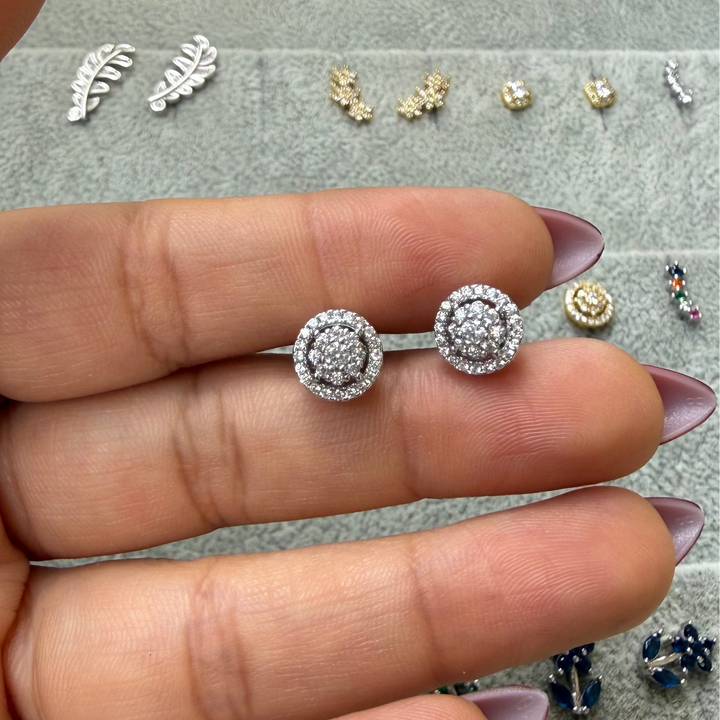 A close-up of a hand holding a pair of silver-toned double oval stud earrings with clear stones, set against a gray jewelry display tray with other earrings in the background.