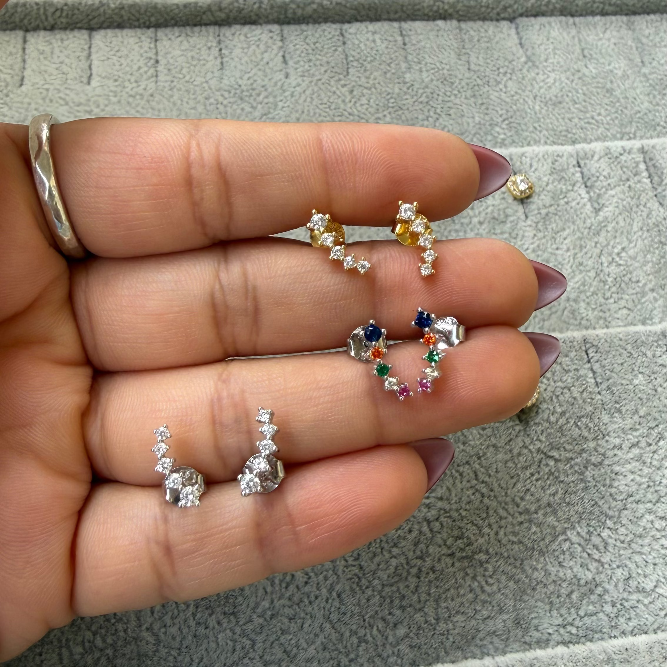 A hand holding several small stud earrings and a matching tray below displays more earrings, including floral and heart-shaped designs in silver, green, blue, and yellow tones.