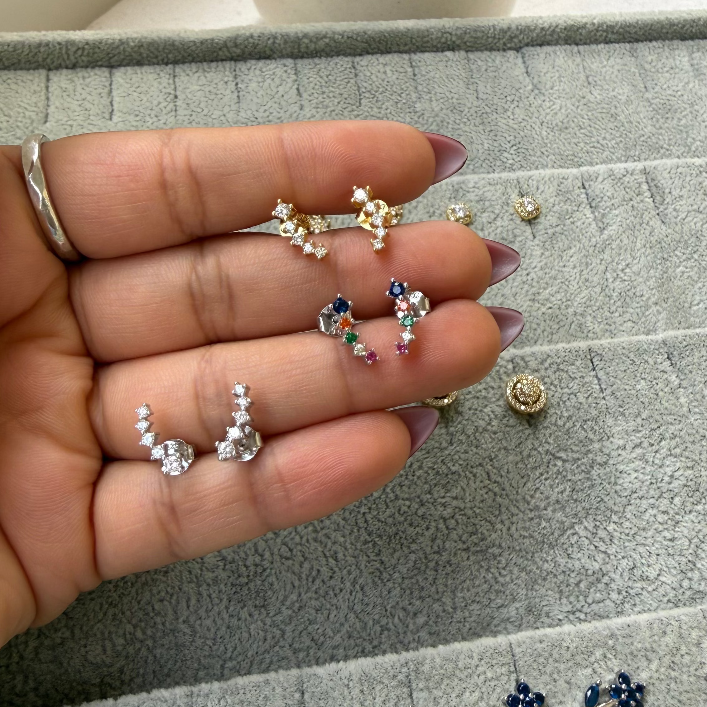 A hand holding several small stud earrings above a gray jewelry tray. The earrings include gold, silver, and multicolor floral and heart designs, with more matching earrings laid out on the tray below.