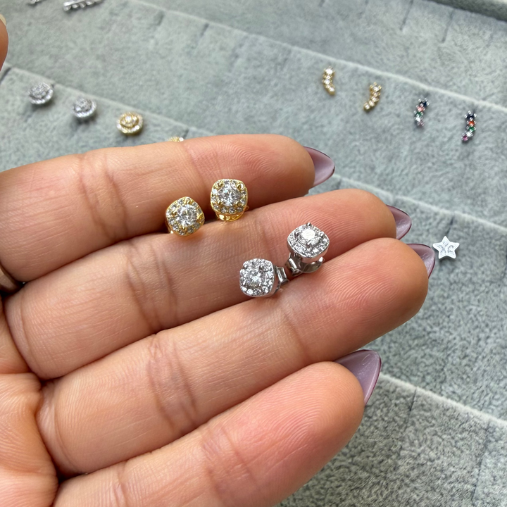 A close-up of a hand holding three small stud earrings in front of a gray jewelry display tray. The earrings appear to be gold and silver-toned with clear gemstones, and other jewelry pieces are visible on the tray in the background.
