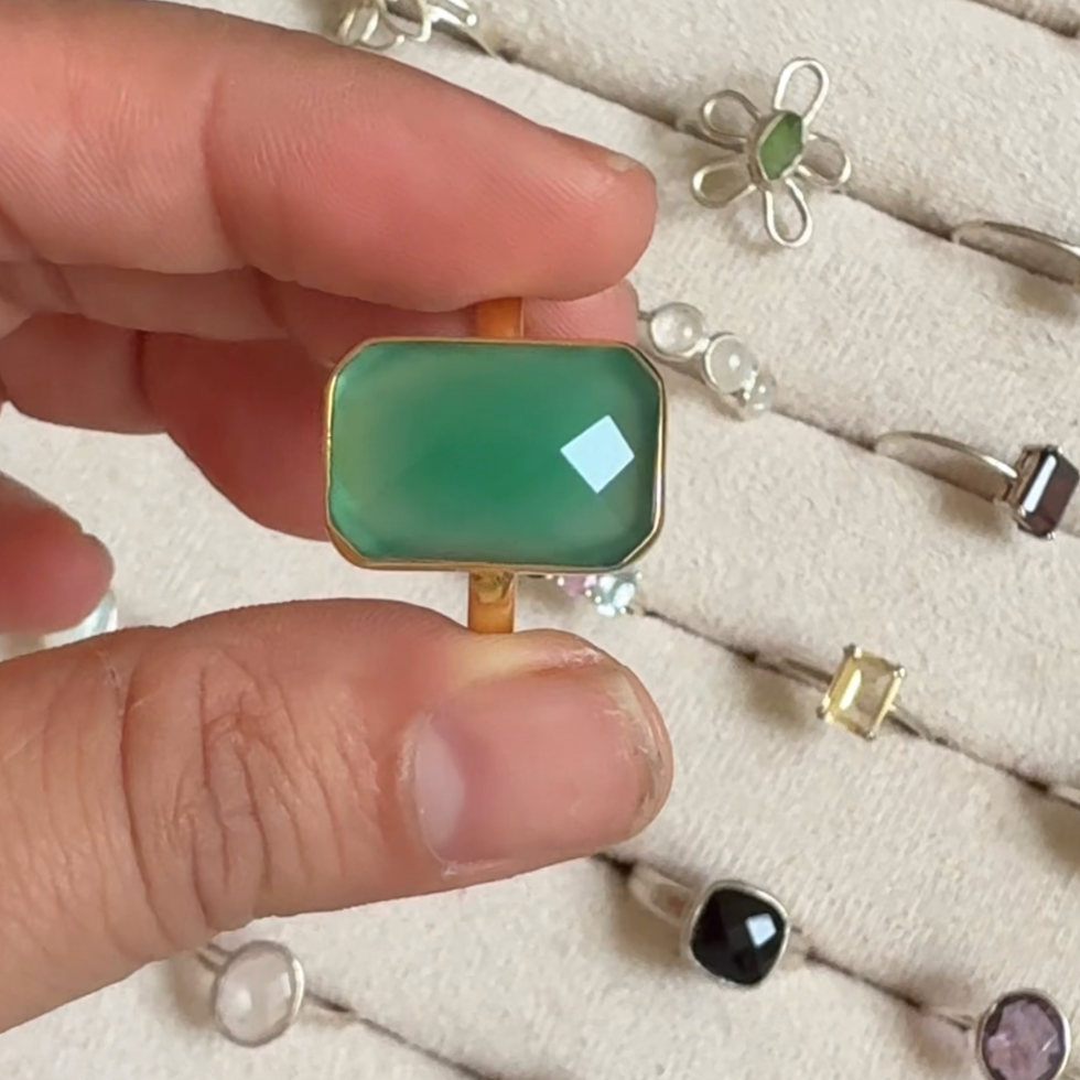 A hand holds a green rectangular ring with a gold band in front of a display of silver rings on a beige jewelry tray. The photo is a screenshot with a video player interface visible, including text overlay and control bars.