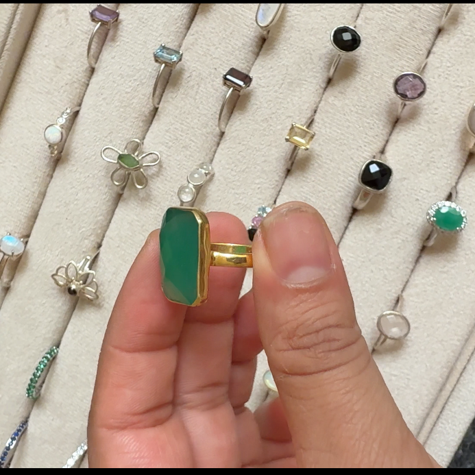 A hand holding a gold ring with a green rectangular stone, shown in a phone app screenshot over a display of multiple rings on a beige tray.