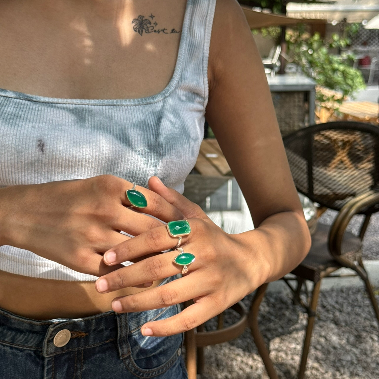 A close-up lifestyle shot of a person wearing a light blue tie-dye camisole and denim jeans, showcasing green rectangular emerald-cut gemstones set in silver rings on their fingers. The background is a blurred outdoor patio setting.