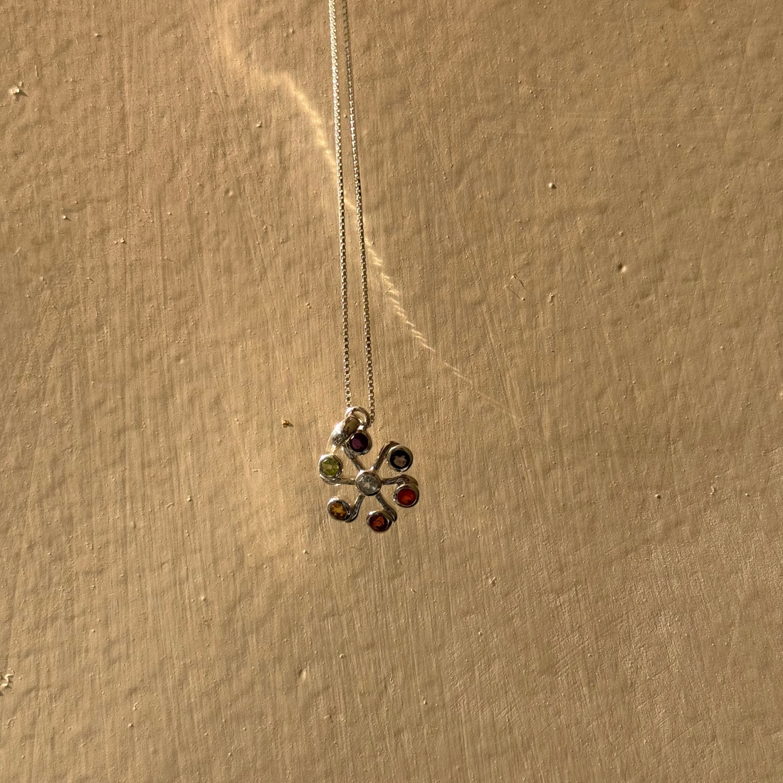 A silver-toned flower-shaped pendant necklace with multiple small colored gemstones, hanging against a textured beige wall.