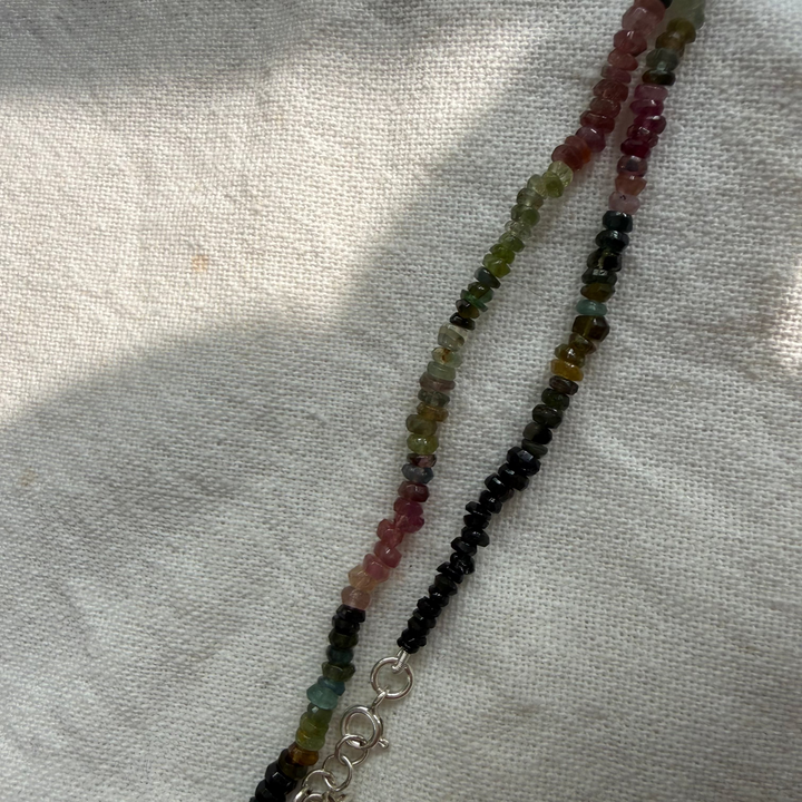 A beaded necklace with alternating strands of black and multicolor round beads, featuring a silver-toned lobster clasp, laid out on a textured off-white fabric.