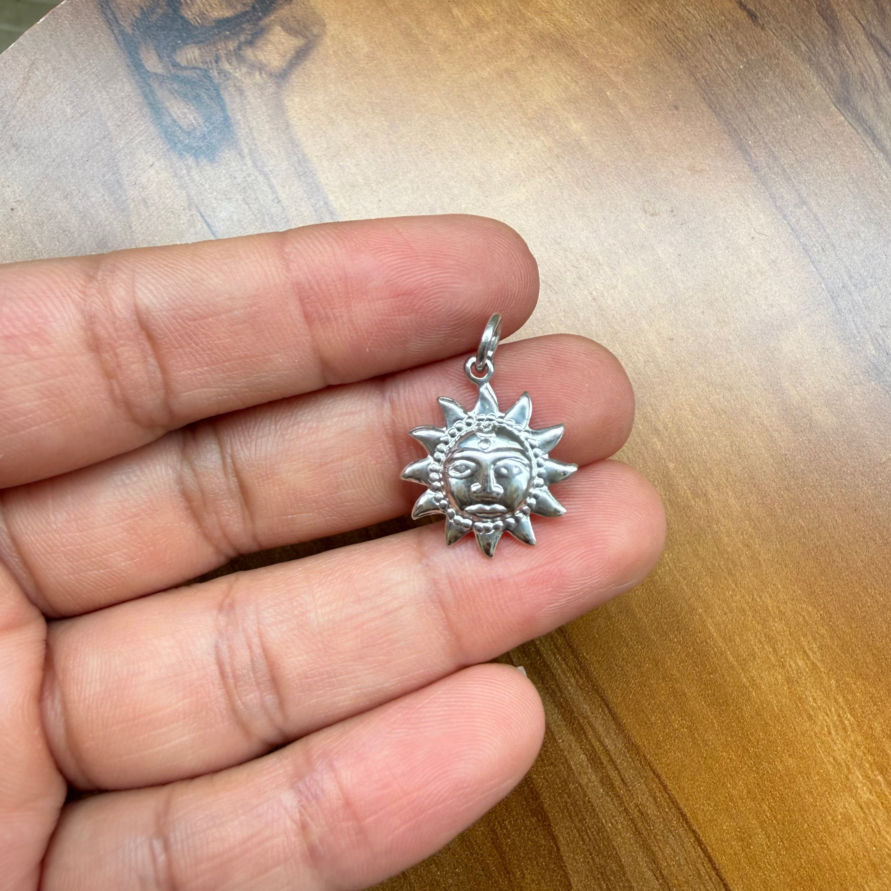 A silver sun pendant with a face in the center, held on a person's fingers over a wooden table.